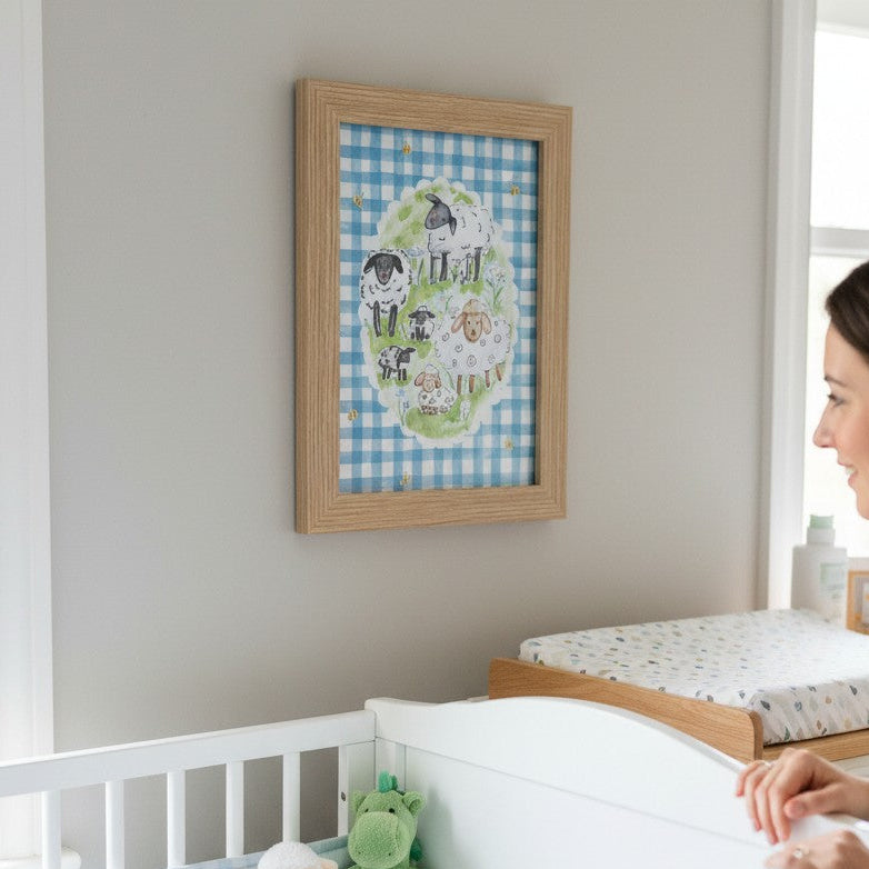 School Bus Studio A4 sheep print in an oak frame hanging in a nursery with blue gingham decor and a baby in a crib.